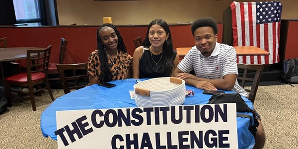 Three students sit behind a table with a blue tablecloth and a sign reading "The Constitution Challenge," with an American flag displayed in the background.   